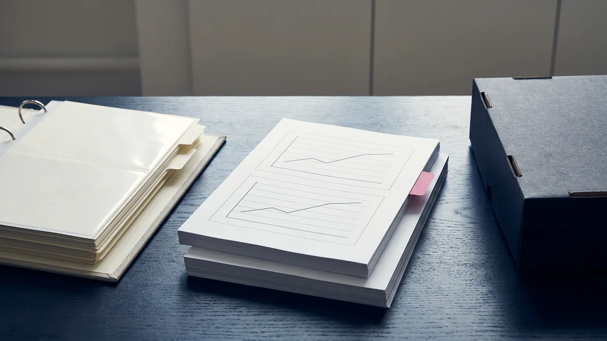 Two offset printed copies of the same chart report with a pink index tab, beside a tabbed binder and a closed archive box on a planning desk.